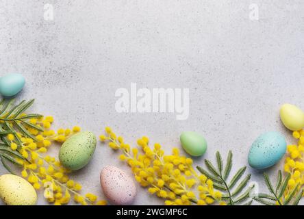 Dieses Bild zeigt ein festliches Osterarrangement aus pastellfarbenen Eiern und hellgelben Mimosa-Blüten auf einer strukturierten hellgrauen Oberfläche. Stockfoto