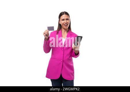 Die junge hübsche Frau mit schwarzen, glatten Haaren trägt eine trendige pinkfarbene Jacke, die eine Kreditkarte und ein Smartphone hält Stockfoto