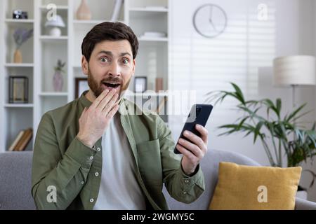 Der verblüffte kaukasier hält das Smartphone in der Hand und bedeckt den Mund mit der Hand, während er die Kamera mit Staunen betrachtet. Beeindruckender Mann, der Reaktion auf unerwartete Textnachrichten auf einem Mobilgerät demonstriert. Stockfoto