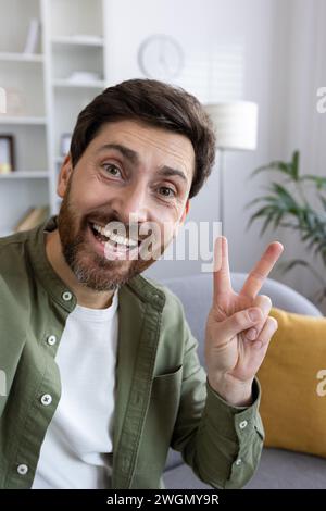 Porträt eines glücklichen bärtigen Mannes, der ein Friedenszeichen mit zwei Fingern über dem Hintergrund des Wohnhauses macht. Gutaussehender Kerl, der Selfie auf der Frontkamera des Smartphones macht oder Videoanrufe startet und einen Freund begrüßt. Stockfoto