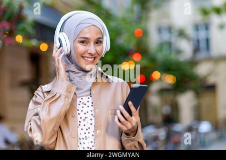 Lächelnde arabische Frau im Hijab hört Hörbuch im Headset, das mit dem Mobilgerät verbunden ist, während sie die Straße hinuntergeht. Attraktive Dame, die Freizeit genießt und moderne Technologie auf unscharfem Hintergrund nutzt. Stockfoto