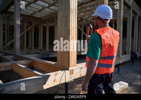 Zimmermann baut ein zweistöckiges Holzrahmenhaus in der Nähe des Waldes. Rückansicht eines Mannes, der Nägel in die Struktur einschlägt, während er Schutzhelm und Bauweste trägt. Stockfoto