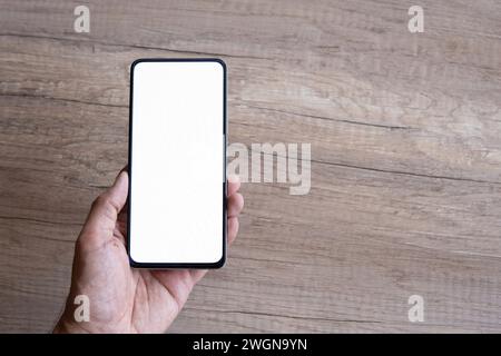 Die Hand des Mannes hält das schwarze Smartphone auf einem Holztisch mit leerem weißen Bildschirm für das Modell. Kopierbereich für Text. Stockfoto