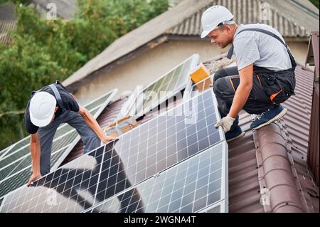 Männer Techniker montieren Photovoltaik-Solarmodule auf dem Dach des Hauses. Elektriker in Helmen, die Sonnenkollektoren im Freien installieren. Konzept alternativer und erneuerbarer Energien. Stockfoto