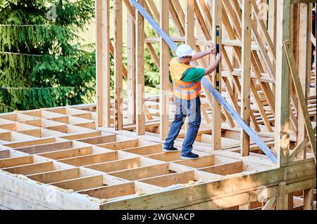 Zimmermann, der ein zweistöckiges Holzrahmenhaus baut. Rückansicht des Mannes überprüft Wände mit Wasserwaage auf Ebenheit und trägt Schutzanzüge, Helm und Weste. Konzept des modernen Bauens. Stockfoto