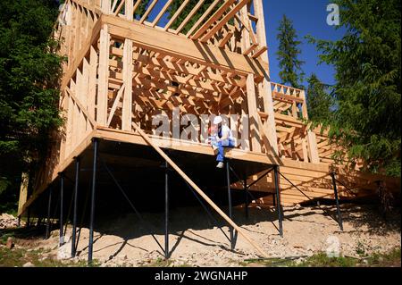 Vater mit Kleinkind-Sohn, der ein Holzrahmenhaus baut. Männlicher Arbeiter, der auf der Baustelle am Balkonrand spielt, Helme und blaue Overalls an sonnigen Tagen trägt. Zimmerei und Familienkonzept. Stockfoto