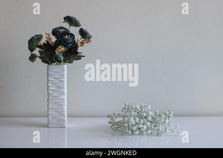 Ein Blumenstrauß aus künstlichen blauen Blumen steht in einer Relief-Vase. Kleine weiße Blumen liegen in der Nähe. Neutraler Hintergrund und Oberfläche Stockfoto