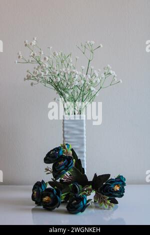 Eine Vase mit Relief-Textur steht auf einer weißen Oberfläche vor einem neutralen Hintergrund. Weiße und blaue Kunstblumen in Blumensträußen Stockfoto