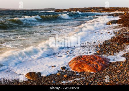 Die dynamische Szene fängt die Energie der Wellen ein, die auf einen Kiesstrand treffen, während die Sonne untergeht und den Himmel mit warmen Farben übermalt. Stockfoto