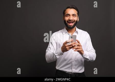 Ein indischer Geschäftsmann in einem weißen Hemd strahlt vor Freude, während er sein Smartphone benutzt, was auf positive Nachrichten oder eine erfolgreiche Transaktion hinweist, vor einem dunkelgrauen Hintergrund Stockfoto