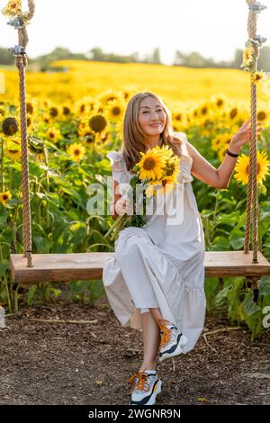 Fröhliche asiatische Frau, die in einem weißen Kleid mit einem Blumenstrauß auf der Schaukel sitzt und Selfie im Sonnenblumenfeld in Yorkshire macht Stockfoto
