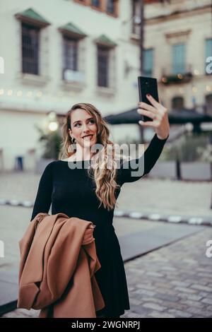 Eine modische Kaukasierin, die ein Selfie auf einer Stadtstraße macht, in einem schwarzen Kleid Stockfoto
