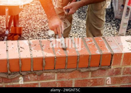 Nahaufnahme einer Mauer aus Ziegelstein und einer Kelle, die der Arbeiter zum Auftragen und Glätten des Mörtels zwischen Ziegelsteinen verwendet. Maurer, der den letzten Schliff auf den Ziegelstein legt Stockfoto