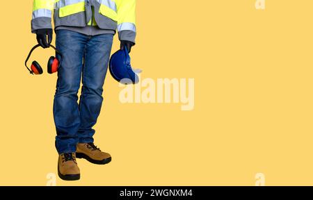 Arbeiter trägt Jeans, Handschuhe, Stiefel und hält Schutzausrüstung, Helm und Ohrenschützer auf gelbem Hintergrund Stockfoto