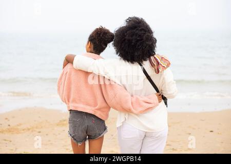 Zwei gemischte Rassenschwestern am Strand umarmen sich und blicken aufs Meer. Stockfoto