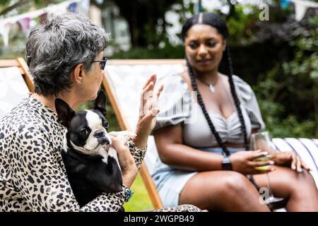 Reife weiße Frau mit einem Boston Terrierhund unterhält sich mit einer jungen schwarzen Frau auf einer Gartenparty im Sommer Stockfoto
