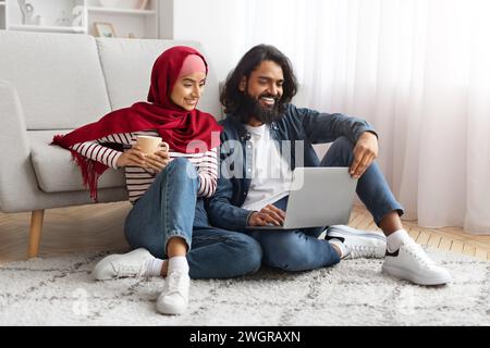 Entspanntes, junges muslimisches Paar, das mit Laptop und Kaffeetasse auf dem Boden sitzt Stockfoto