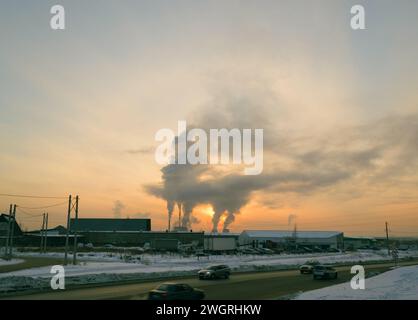 Verkehr von Autos auf einer Vorortstraße vor dem Hintergrund des Rauches aus einem Wärmekraftwerk und vor dem Hintergrund des Abendhimmels bei Sonnen Stockfoto