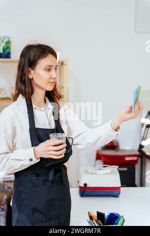 Buntglaskünstler arbeiten im Studio. Eine junge Frau in Schürze wählt Glas für ihre zukünftige Arbeit. Vertikale Aufnahme. Hobbys, Kunst und Kunsthandwerk. Stockfoto