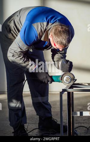 Ein Facharbeiter, der sich auf das Schleifen eines Metallstücks mit einem tragbaren Elektrowerkzeug in einer hell beleuchteten industriellen Umgebung konzentriert, wird gezeigt. Stockfoto