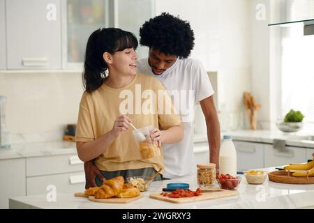 Eine junge lächelnde Frau, die ihren afroamerikanischen Ehemann ansah, während sie in der Küche am Tisch stand und das Frühstück zubereitete Stockfoto