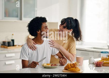Eine junge lächelnde Frau, die ihren glücklichen Mann ansieht, der in der Küche am Tisch sitzt und frühstückt, während er ihn umschließt Stockfoto