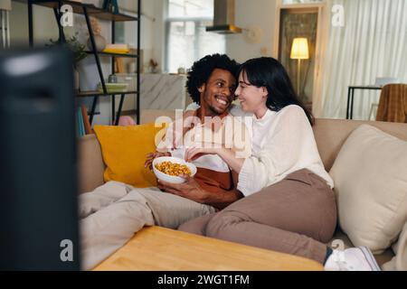 Junge liebevolle Mann und Frau, die sich anschauen und Popcorn essen, während sie zu Hause auf der Couch vor dem Fernseher sitzen Stockfoto