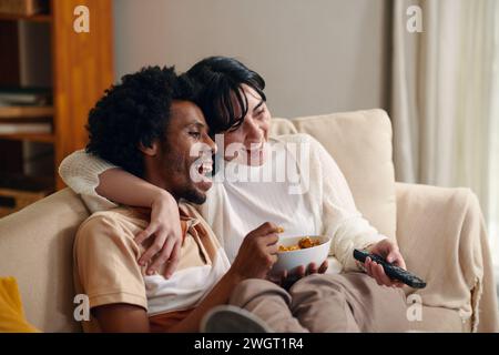 Junger lachender Mann mit einer Schale Popcorn und seine Frau mit Fernbedienung sitzt auf der Couch vor dem Fernseher und guckt Sitcom Stockfoto
