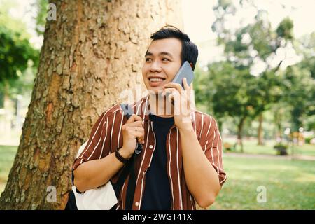 Lächelnder asiatischer Kerl mit Rucksack, der im Park gegen Baumstamm steht und mit einem Freund auf dem Smartphone spricht, während er beiseite blickt Stockfoto