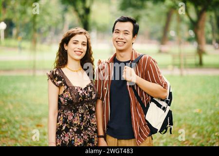 Glückliches junges interkulturelles Paar in Casualwear, das im Sommer auf dem Campus oder im großen Park mit einem Lächeln in die Kamera blickt Stockfoto
