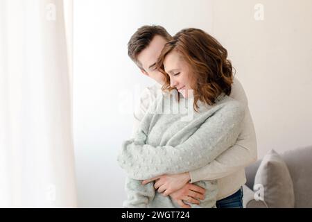 Liebevoller junger Mann, der die Frau umschließt und sich von hinten in einem hellen Wohnzimmer zu Hause küsst. Romantisches glückliches Paar in legeren Kleidern, das eine gute Zeit verbringt Stockfoto