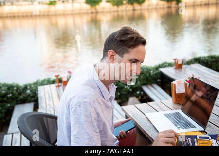 Der junge Mann schaut sich die Speisekarte an, er arbeitet an seinem Laptop in einem Restaurant am Fluss Stockfoto