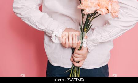 Ein Junge in einem weißen Hemd hält einen Blumenstrauß hinter dem Rücken vor einem rosa Hintergrund Stockfoto