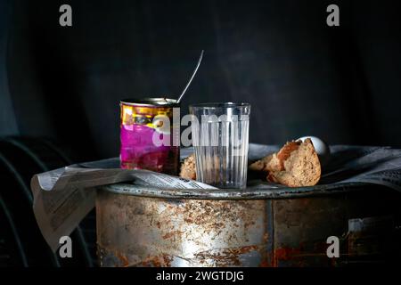 Bild eines leeren Glases und Brotstückchen in der Nähe einer offenen Dose auf einem rostigen Fass Stockfoto