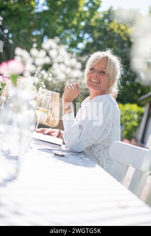 Attraktive blonde Mutter sitzt an ihrem Gartentisch in der Sonne lachend und entspannend. Verschwommener Garten und Blumen im Hintergrund. Ein Glas Wein auf dem Tisch Stockfoto