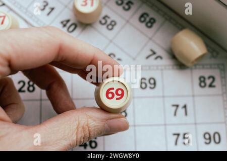 Lottospiel spielen. Person, die den Würfel mit der Figur auf dem Hintergrund der Bingo-Karte hält. Nostalgie Lifestyle. Tischspiele. Retro-Spiele. Zylinder mit der Nummer 69. Stockfoto