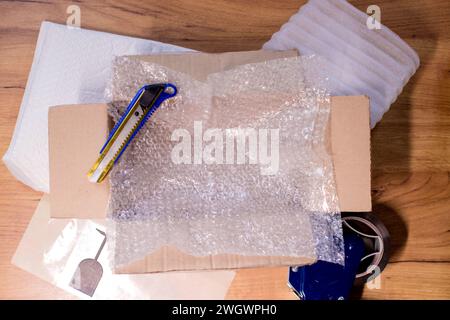 Offene Schachtel mit verpackten Gegenständen, Klebeband, Schere, Papier und Luftpolsterfolie auf Holztisch, flach gelegt. Stockfoto