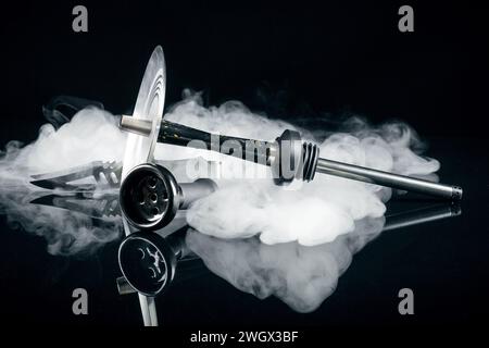 Shisha mit Schüssel und Zange auf schwarzem Hintergrund. Künstlerische Aufnahmen in einem Atelier mit weißem Rauch und Reflexion. Stockfoto