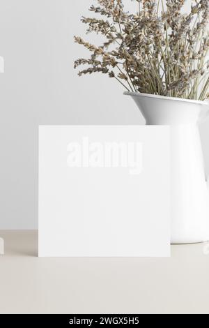 Quadratische Einladungskarte Mockup mit getrocknetem Lavendel auf einem beigen Tisch. Stockfoto