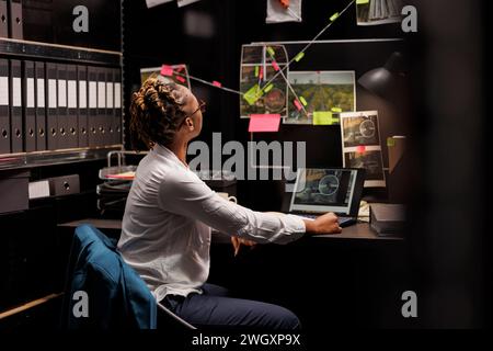 Die afroamerikanische Polizistin analysiert die Beweisstelle, während sie Verbrechen aufklärt. Strafverfolgungsexperte, die sich Hinweise ansehen, die an der Wand hängen, und Einblicke suchen Stockfoto