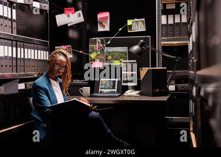 afroamerikanerin sitzt am Schreibtisch des Detektivs und liest die Akte des Verbrechens. Privatdetektiv, der forensische Inspektorenschlussfolgerungen studiert und Nachtansicht sucht Stockfoto