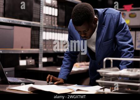 Polizist liest Kriminologiebericht, arbeitet spät in der Nacht in einem Kriminalfall im Arhive Room. afroamerikanischer Ermittler analysiert Tatortbeweise und sucht nach neuen Hinweisen Stockfoto