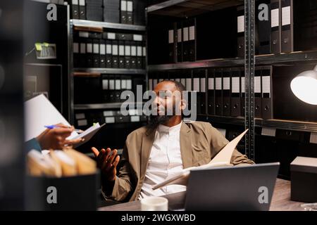 Detective bespricht Ideen zur Kriminalauflösung mit einem Assistenten im Büro nachts. afroamerikaner-Ermittler spricht über Beweisanalyse, während eine Kollegin Notizen macht Stockfoto