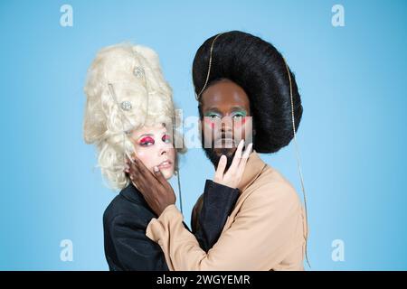 Porträt von schwulen männlichen und weiblichen Models mit Make-up und Vintage Perücken. Ernsthafte LGBT-Menschen, die sich gegenseitig umarmen und berühren und in die Kamera schauen. Paare posieren auf blauem Hintergrund. Stockfoto