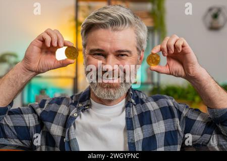 Lächelnder Kaukasischer Senior Freelancer man Aktienhändler hält goldene Bitcoins in der Nähe des Gesichts. Glücklicher aufgeregter männlicher Mann mittleren Alters erfolgreicher Entwickler-Programmierer im Wohnzimmer zu Hause. Steigerung des Wohlstands Stockfoto