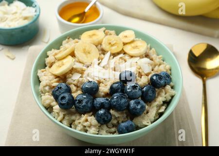 Leckeres Haferbrei mit Banane, Heidelbeeren, Kokosflocken und Honig, serviert in einer Schüssel auf beigem Tisch Stockfoto