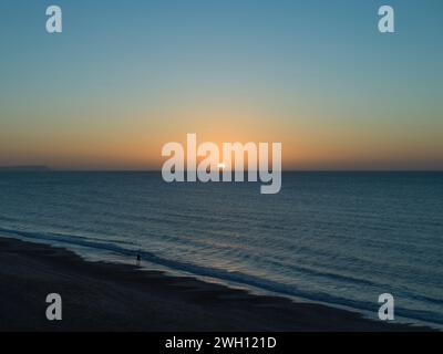Ein einsamer Mann läuft an einem Wintermorgen bei Sonnenaufgang an der Flutlinie entlang. Es gibt keine Wolken am blauen und orangen Sonnenaufgangshimmel im Januar und das Meer ist ruhig. Stockfoto