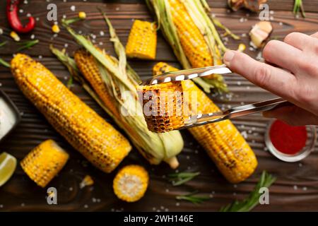 Küchenzange mit gegrilltem Mais auf den Kolbenstücken in kaukasischen Händen auf Küche Holztisch Hintergrund Stockfoto