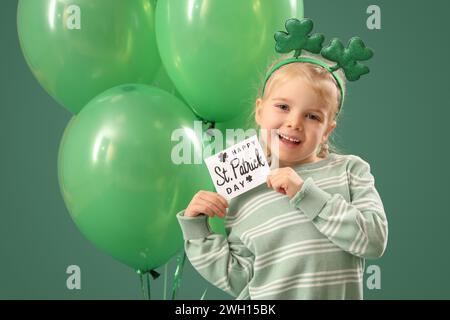 Niedliches kleines Mädchen mit Leprechaun Hut, Grußkarte und Ballons auf grünem Hintergrund. St. Patrick's Day-Feier Stockfoto