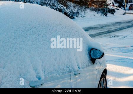 Geparktes weißes Auto bedeckt mit einer dicken Schneeschicht, bedeckt Oberseite, Türen und Fenster, gefrorene Straße und Büsche im Hintergrund verschwimmen, Wintertag Stockfoto